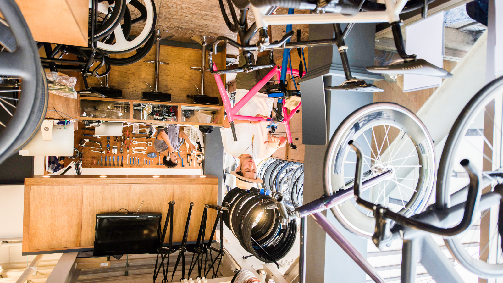 Two employees working in a bicycle repair shop with bikes and tools in the background. One puts together a bicycle and the other is on the phone behind the repair counter.